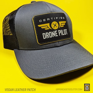 May include: A gray and black trucker hat with a black vegan leather patch that says "CERTIFIED DRONE PILOT" in white text with a yellow drone icon and yellow wings.