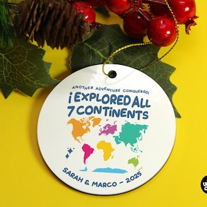 May include: A white round Christmas ornament with a world map design in various colors. The text reads "¡ EXPLORED ALL 7 CONTINENTS" with the names "SARAH & MARCO - 2025" below. The ornament is hanging with a gold string.