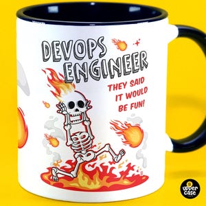 Puede incluir: Taza de café de cerámica blanca con borde y asa negros. La taza presenta un esqueleto de dibujos animados en llamas con el texto "DevOps Engineer" y "¡Dijeron que sería divertido!"