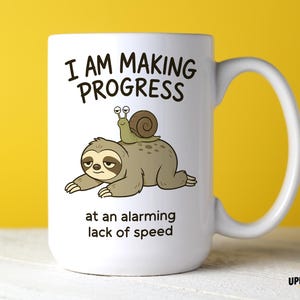 May include: A white ceramic mug featuring a cartoon illustration of a sloth with a snail on its back. The text reads I AM MAKING PROGRESS at an alarming lack of speed. The mug has a standard handle and a smooth, cylindrical body design.