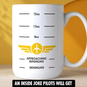 May include: A white ceramic mug featuring black measurement lines for 8oz, 12oz, and 15oz. Below the lines is a yellow airplane icon with wings, followed by the black text "APPROACHING MINIMUMS" and "MINIMUMS" printed on the front of the mug.