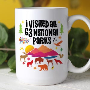 May include: A white ceramic mug featuring the black text I VISITED ALL 63 NATIONAL PARKS, a colorful map of the United States, a sunset over mountains, and various wildlife illustrations including a moose, bear, wolf, bison, and eagle.