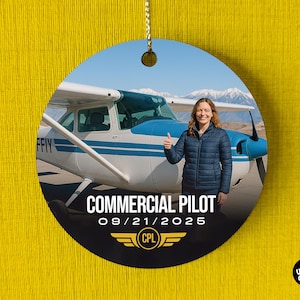 Puede incluir: Adorno redondo con una mujer haciendo una señal de pulgar hacia arriba frente a un avión pequeño. El adorno tiene el texto "COMMERCIAL PILOT" y la fecha "09/21/2025" con un logotipo CPL. El fondo muestra montañas y cielo azul.