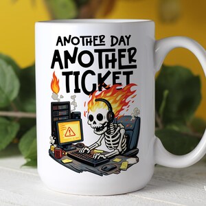 Puede incluir: Taza de cerámica blanca con un diseño humorístico. La taza presenta un esqueleto con llamas como pelo, con auriculares y trabajando en una computadora. El texto dice "ANOTHER DAY ANOTHER TICKET".