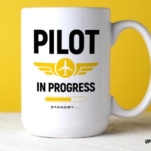 Könnte beinhalten: Ein weißer Keramikbecher mit dem schwarzen Text PILOT IN PROGRESS und einem gelben Flugzeugsymbol zwischen den Wörtern. Unter dem Text befindet sich eine gelbe Ladebalkengrafik und das Wort STANDBY in kleineren schwarzen Großbuchstaben.
