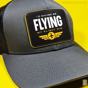 Puede incluir: Una gorra de camionero gris y negra con un parche amarillo y negro que dice "I'd rather be flying my flight sim". El parche presenta un avión amarillo con alas.