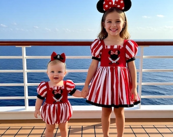 May include: Two children wearing matching red and white striped dresses with black accents and Minnie Mouse-themed headbands. The dresses feature a Minnie Mouse design on the bodice. They are standing on a wooden deck with a blue ocean in the background.