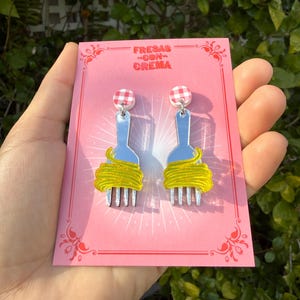 May include: Novelty earrings with a fork design, yellow spaghetti, and red and white gingham studs. The earrings are presented on a pink card with the text "Fresas Con Crema".