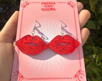 May include: Red acrylic lip-shaped earrings with a mosaic-like design, hanging from silver hooks. The earrings are displayed on a pink card with the text "Fresas con Crema" in a decorative frame. The background is a green, leafy environment.