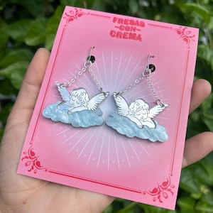 Peut inclure: Une paire de boucles d'oreilles en argent en forme d'ange avec un design de nuage bleu et blanc. Les boucles d'oreilles sont sur une carte rose avec le texte "FRESAS CON CREMA".