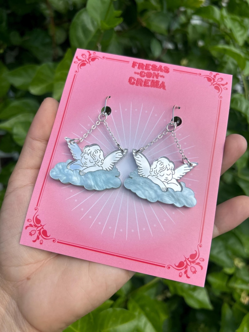 Peut inclure: Une paire de boucles d'oreilles en argent en forme d'ange avec un design de nuage bleu et blanc. Les boucles d'oreilles sont sur une carte rose avec le texte "FRESAS CON CREMA".