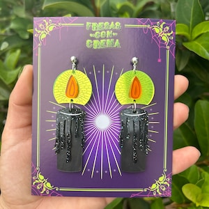 May include: A pair of black candle earrings with dripping wax and glitter accents. The earrings are on a purple card with the text "FRESAS CON CREMA".