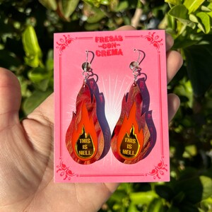 May include: Flame-shaped earrings in red and orange hues, with the text "THIS IS HELL" in gold. The earrings are presented on a pink card, which also features the text "FRESAS CON CREMA".