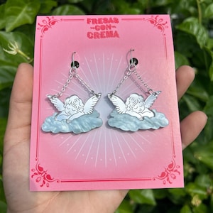 Peut inclure: Une paire de boucles d'oreilles en argent en forme d'ange avec un design de nuage bleu et blanc. Les boucles d'oreilles sont sur une carte rose avec le texte "FRESAS CON CREMA".