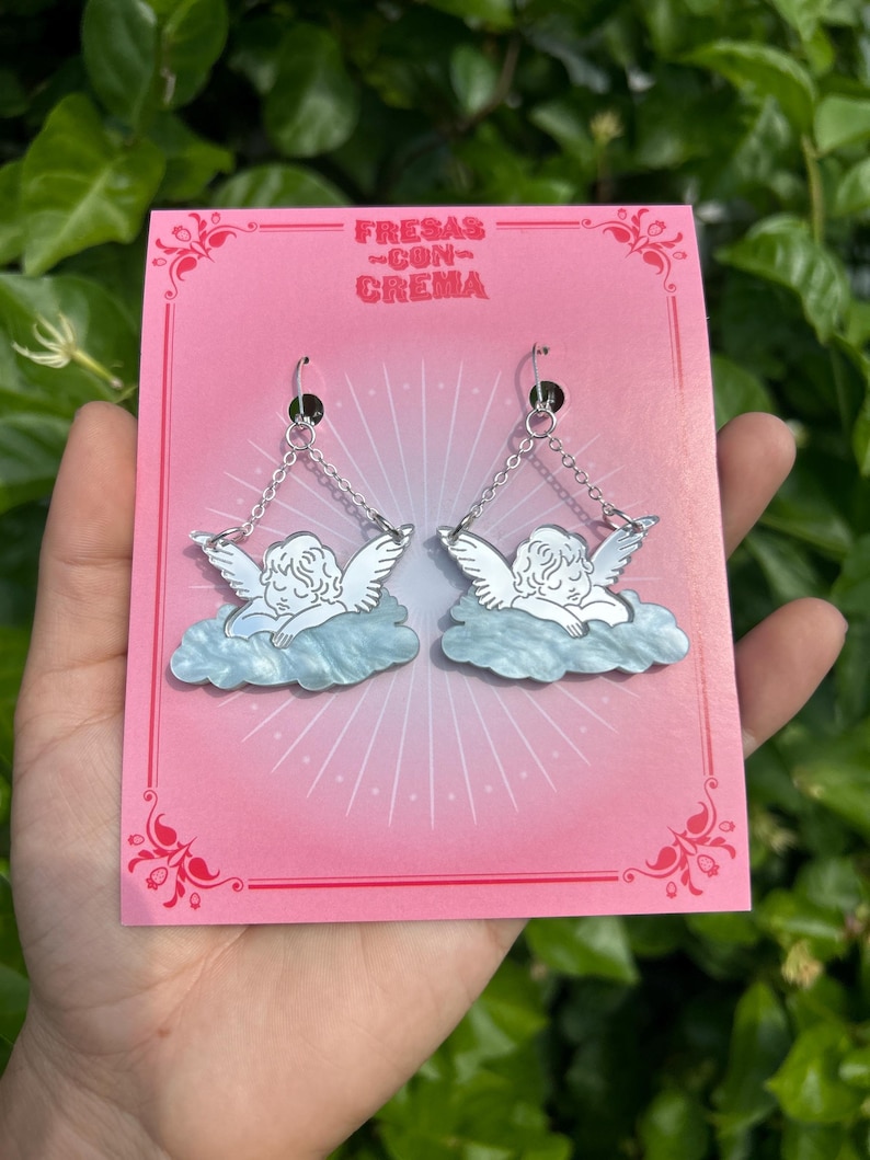 Peut inclure: Une paire de boucles d'oreilles en argent en forme d'ange avec un design de nuage bleu et blanc. Les boucles d'oreilles sont sur une carte rose avec le texte "FRESAS CON CREMA".