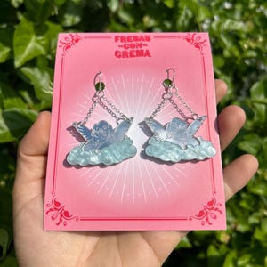 Peut inclure: Une paire de boucles d'oreilles pendantes en argent en forme d'ange avec un design de nuage bleu et blanc. Les boucles d'oreilles sont sur une carte rose avec le texte "FRESAS -CON- CREMA".