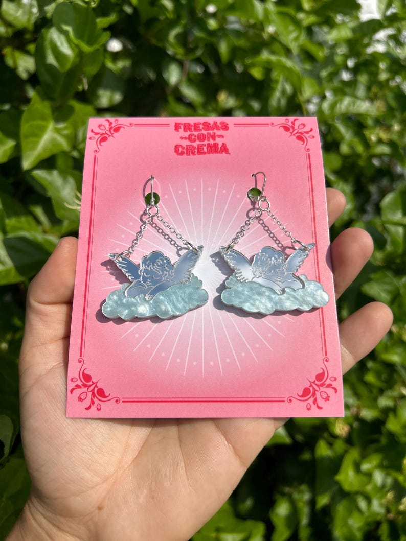 Peut inclure: Une paire de boucles d'oreilles pendantes en argent en forme d'ange avec un design de nuage bleu et blanc. Les boucles d'oreilles sont sur une carte rose avec le texte "FRESAS -CON- CREMA".