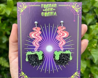 May include: A pair of black cauldron earrings with green glitter slime and pink and white swirl accents. The earrings are on a purple background with the text "FRESAS CON CREMA".