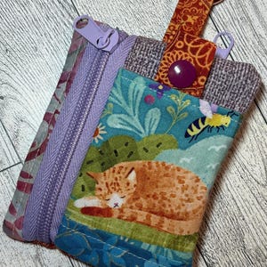 May include: A fabric pouch featuring a sleeping orange tabby cat and a bee on a teal background. The pouch has a lavender zipper, a dark purple snap closure on an orange patterned tab, a copper-colored metal ring, and a gray textured fabric section.