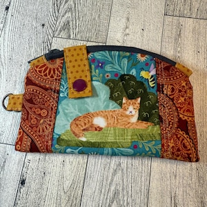 May include: A fabric zipper pouch featuring a central panel with a ginger cat resting on a green surface, surrounded by blue floral patterns and a small bee. The sides are made of a dark orange fabric with a circular paisley-like print.