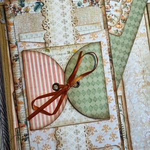 May include: A handmade scrapbook with a fall theme. The scrapbook features patterned paper with floral and geometric designs, a ribbon closure, and a card with a scarecrow illustration. The color palette includes shades of orange, green, and cream.