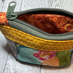 May include: A small fabric zipper pouch featuring a colorful cat print with a floral collar on the front panel. The pouch has a yellow dotted fabric trim, a grey zipper, and an interior lining with a dark orange and brown paisley pattern.