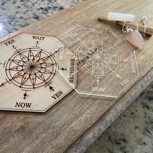 May include: A wooden divination board with a geometric design and the words "Yes," "No," "Maybe," "Wait," and "Now." A clear acrylic board with a similar design is next to it. A rose quartz pendulum and selenite wand are also present.