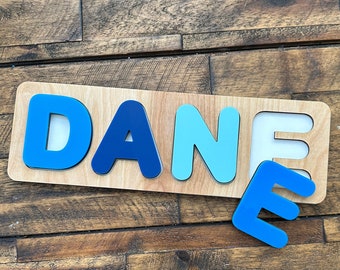 May include: Wooden puzzle spelling the name "DANE" in blue letters. The puzzle has a light wood base with the letters in varying shades of blue. The letter "E" is separate from the rest of the puzzle.