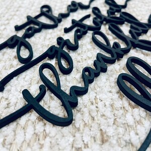 May include: Black laser-cut wooden word cutouts in a cursive font. The words "love", "thank", and "grateful" are visible. The cutouts are scattered on a textured, off-white surface. Ideal for crafts and DIY projects.