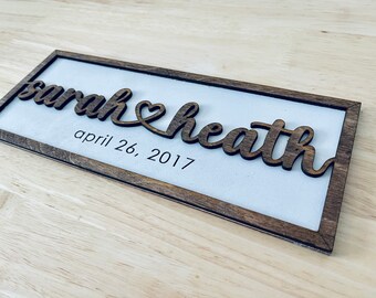 May include: Wooden sign with the names "Sarah & Heath" in a cursive font, joined by a heart symbol. The date "april 26, 2017" is printed below the names. The sign has a white background and a dark brown wooden frame.