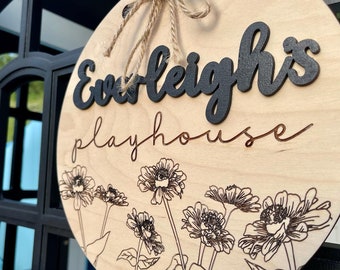 May include: A circular wooden sign, reading "Everleigh's Playhouse" in black and brown script. The sign has a floral design at the bottom and is suspended by a twine rope. The sign is made of light-coloured wood.