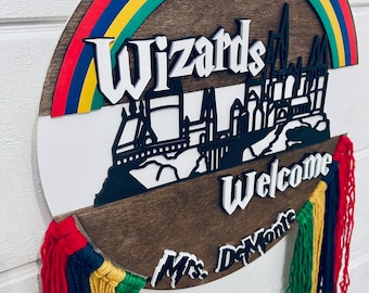 May include: A round wooden sign with a rainbow arch and the words "Wizards Welcome" in white. The sign features a silhouette of a castle and colorful yarn tassels in red, green, yellow, and blue. The name "Mrs. DeMonte" is also displayed.