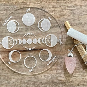 May include: A clear acrylic moon phase board with etched moon cycle designs and the words "yes", "no", "maybe", and "unknown". A rose quartz pendulum and a selenite wand rest beside the board, along with a piece of Palo Santo wood.