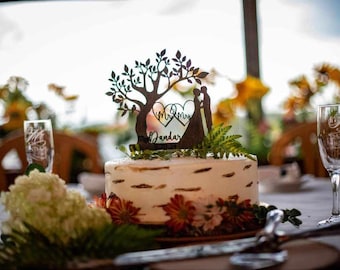 May include: A wedding cake with a tree-shaped topper, a bride and groom silhouette, and the words 'Mr & Mrs'. The cake is decorated with flowers and greenery. Champagne flutes are in the background.