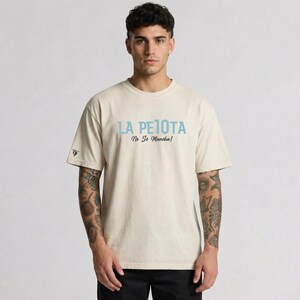 Puede incluir: Una camiseta beige con el texto "LA PELOTA No Se Mancha!" en azul claro. La camiseta tiene mangas cortas y un peque&ntilde;o logo en la manga izquierda. La persona lleva pantalones negros.