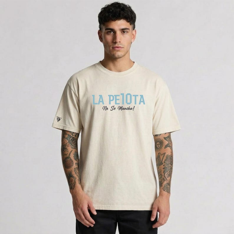 Puede incluir: Una camiseta beige con el texto "LA PELOTA No Se Mancha!" en azul claro. La camiseta tiene mangas cortas y un peque&ntilde;o logo en la manga izquierda. La persona lleva pantalones negros.
