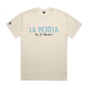 Puede incluir: Camiseta color crema con el texto "LA PELOTA No Se Mancha!" en azul claro. La camiseta tiene cuello redondo y mangas cortas. Un peque&ntilde;o logotipo es visible en la manga izquierda.