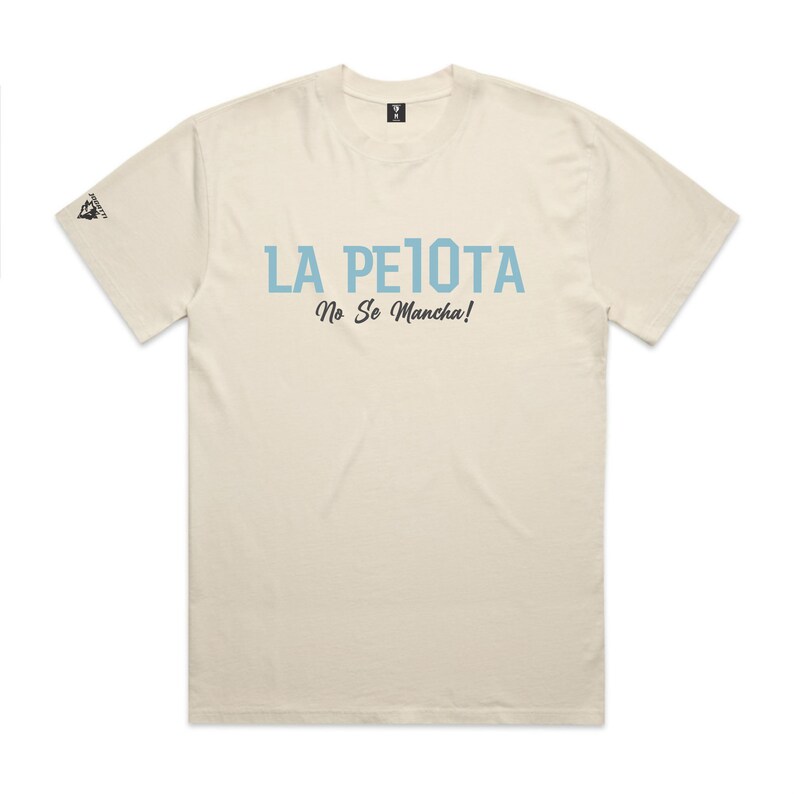 Puede incluir: Camiseta color crema con el texto "LA PELOTA No Se Mancha!" en azul claro. La camiseta tiene cuello redondo y mangas cortas. Un peque&ntilde;o logotipo es visible en la manga izquierda.