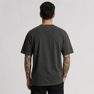 Puede incluir: Una camiseta gris oscuro de manga corta. La parte trasera de la camiseta es visible, y la persona que la lleva tiene tatuajes en los brazos. La camiseta tiene un aspecto ligeramente descolorido y un corte relajado. La tela es suave.
