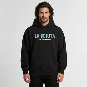 Puede incluir: Sudadera con capucha negra con el texto "LA PELOTA No Se Mancha!" en azul claro. La sudadera tiene un bolsillo delantero y una capucha con cord&oacute;n. La imagen muestra a una persona que lleva la sudadera.