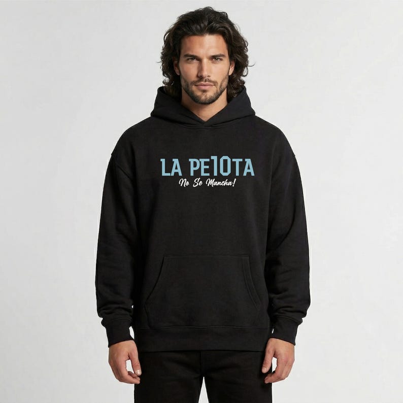 Puede incluir: Sudadera con capucha negra con el texto "LA PELOTA No Se Mancha!" en azul claro. La sudadera tiene un bolsillo delantero y una capucha con cord&oacute;n. La imagen muestra a una persona que lleva la sudadera.