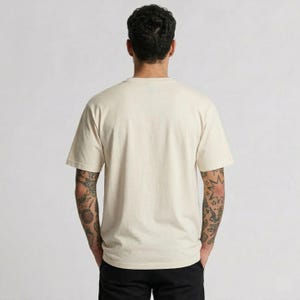Puede incluir: Camiseta de manga corta color crema. La camiseta tiene cuello redondo y un corte relajado. La persona lleva pantalones negros. El fondo es de color gris claro liso. La persona tiene tatuajes en ambos brazos.