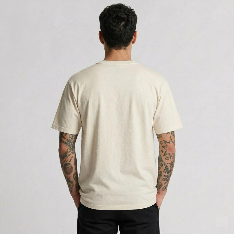 Puede incluir: Camiseta de manga corta color crema. La camiseta tiene cuello redondo y un corte relajado. La persona lleva pantalones negros. El fondo es de color gris claro liso. La persona tiene tatuajes en ambos brazos.