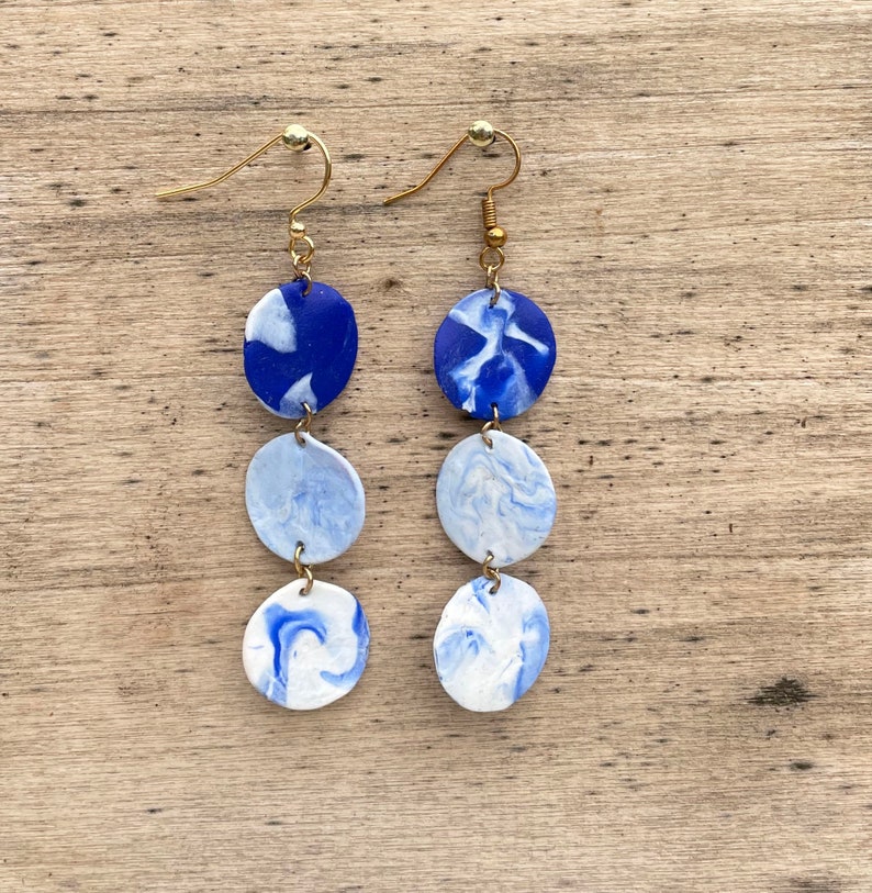 Puede incluir: Pendientes colgantes con dise&ntilde;o veteado. Cada pendiente presenta tres colgantes circulares veteados en tonos azules y blancos, conectados por ganchos y alambres dorados. Los pendientes cuelgan de ganchos para la oreja dorados.