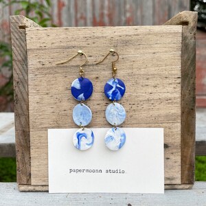 Puede incluir: Pendientes hechos a mano con un dise&ntilde;o azul y blanco veteado. Cada pendiente presenta tres colgantes circulares apilados, suspendidos de alambres dorados. Los pendientes se exhiben sobre una superficie de madera, con el texto "papermoonn studio".