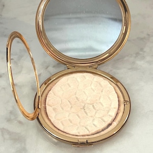 Peut inclure: Un bo&icirc;tier compact rond en m&eacute;tal dor&eacute; avec un couvercle ouvert r&eacute;v&eacute;lant un miroir circulaire et une poudre press&eacute;e. La poudre pr&eacute;sente une texture g&eacute;om&eacute;trique en forme de nid d'abeille sur toute sa surface, ins&eacute;r&eacute;e dans le cadre m&eacute;tallique.