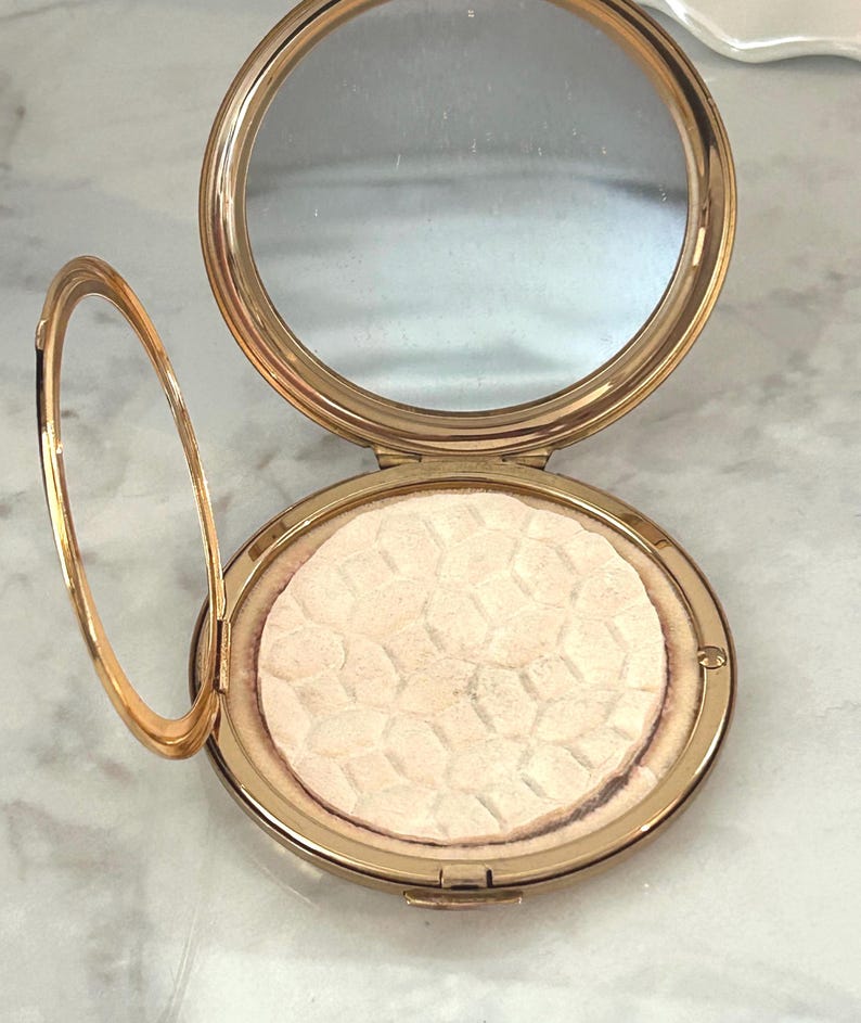 Peut inclure: Un bo&icirc;tier compact rond en m&eacute;tal dor&eacute; avec un couvercle ouvert r&eacute;v&eacute;lant un miroir circulaire et une poudre press&eacute;e. La poudre pr&eacute;sente une texture g&eacute;om&eacute;trique en forme de nid d'abeille sur toute sa surface, ins&eacute;r&eacute;e dans le cadre m&eacute;tallique.