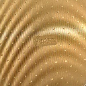 Peut inclure: Un poudrier rond en m&eacute;tal dor&eacute; pr&eacute;sentant une surface textur&eacute;e avec des lignes verticales et de petits motifs en chevron r&eacute;p&eacute;t&eacute;s. Le centre affiche un logo rectangulaire en relief portant l'inscription Stratton et Made in England.