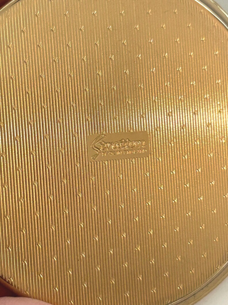 Peut inclure: Un poudrier rond en m&eacute;tal dor&eacute; pr&eacute;sentant une surface textur&eacute;e avec des lignes verticales et de petits motifs en chevron r&eacute;p&eacute;t&eacute;s. Le centre affiche un logo rectangulaire en relief portant l'inscription Stratton et Made in England.