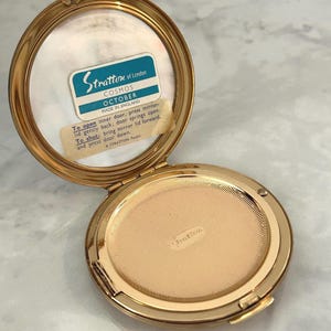 May include: A round gold-tone metal powder compact with an open lid. The interior features a mirror with a label reading Stratton of London COSMOS OCTOBER MADE IN ENGLAND and instructions for use. The base has a textured gold-tone metal surface.